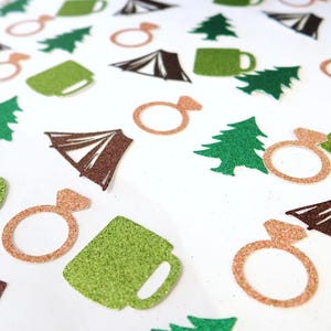 May include: A collection of glittery paper confetti pieces in the shapes of green pine trees, green mugs, brown camping tents, and rose gold engagement rings scattered across a flat surface, creating a festive and thematic decorative arrangement.