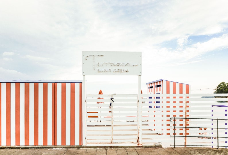 May include: A white gate with a sign that reads "Tamerici Bagni e Spiaggia" above it. The gate is surrounded by a white fence with red and white stripes.