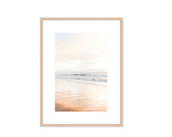 May include: A framed photograph of a beach scene with soft, golden sand and gentle waves lapping at the shore. The sky is a pale blue with wispy clouds, and the sun is shining brightly.