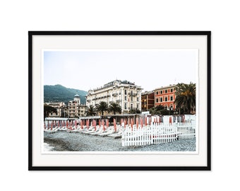 May include: A photograph of a beach scene with a white picket fence, red and white beach umbrellas, and a row of white lounge chairs. The beach is lined with buildings, including a large, white building with a red roof.