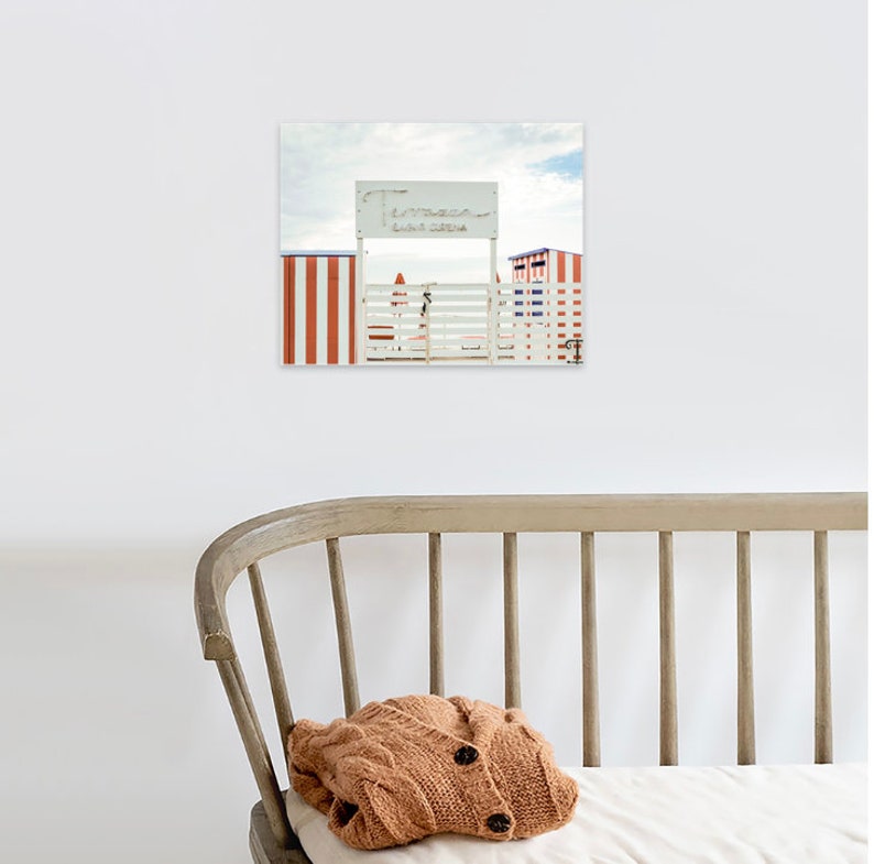 May include: A white wooden gate with a sign that reads "Lido di Venezia" in front of red and white striped beach huts.