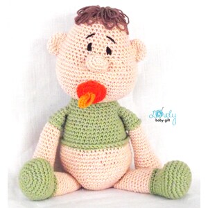 May include: A handmade crochet baby doll with brown hair, a pacifier, and a green shirt. The doll is wearing a diaper and has green feet. The doll is sitting down and has a neutral expression. The doll is a baby gift.