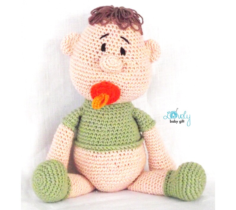 May include: A handmade crochet baby doll with brown hair, a pacifier, and a green shirt. The doll is wearing a diaper and has green feet. The doll is sitting down and has a neutral expression. The doll is a baby gift.