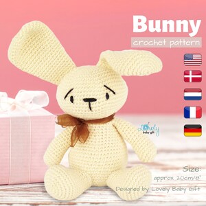Peut inclure: Un modèle de lapin en crochet de couleur crème avec un ruban brun. Le lapin est assis sur une surface en bois. Le modèle est disponible en plusieurs langues, dont l'anglais, le danois, le néerlandais, le français et l'allemand. Le lapin mesure environ 20 cm de haut.