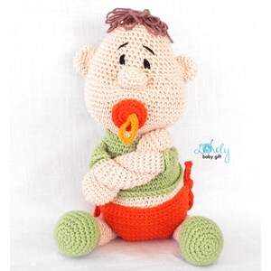 May include: A handmade crochet baby doll with a pacifier. The doll has a light skin tone, brown hair, and is wearing a green shirt and orange diaper. The doll is sitting with its arms crossed.