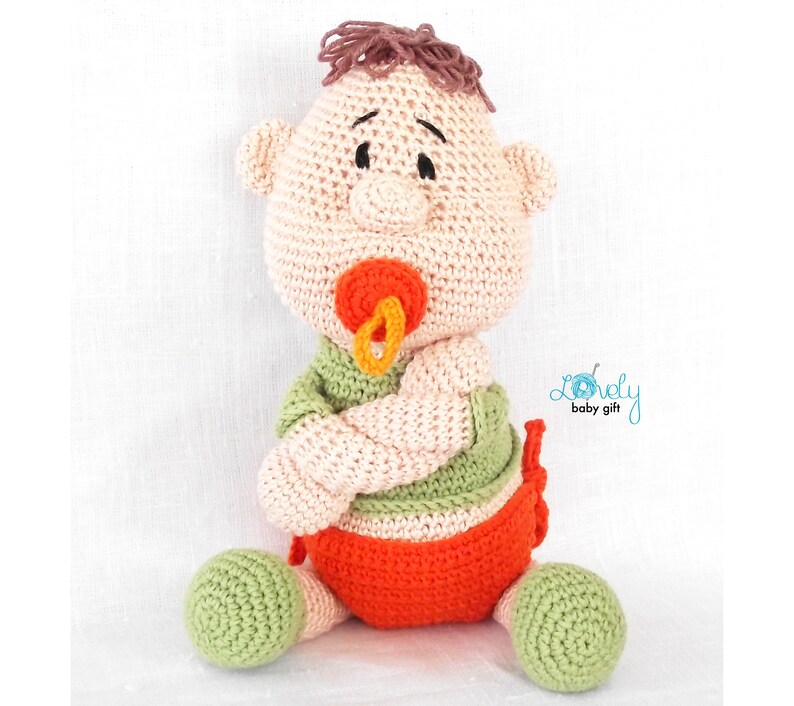 May include: A handmade crochet baby doll with a pacifier. The doll has a light skin tone, brown hair, and is wearing a green shirt and orange diaper. The doll is sitting with its arms crossed.