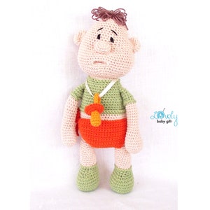 May include: A handmade crochet doll in the shape of a baby. The doll has a light skin tone, brown hair, and is wearing a green shirt, orange diaper, and green shoes. A pacifier hangs around its neck. The doll is a baby gift.