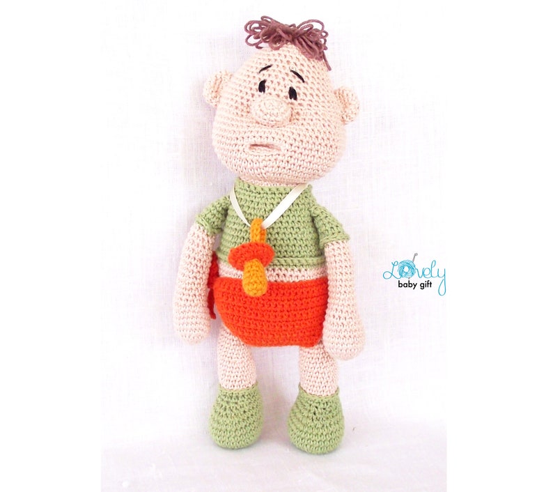 May include: A handmade crochet doll in the shape of a baby. The doll has a light skin tone, brown hair, and is wearing a green shirt, orange diaper, and green shoes. A pacifier hangs around its neck. The doll is a baby gift.