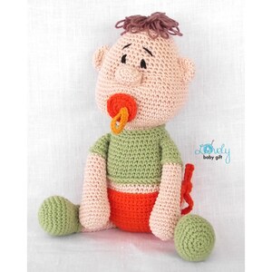 May include: A handmade crochet baby doll with a pacifier. The doll has a light skin tone, green shirt, orange shorts, and green feet. The doll's hair is brown, and it has a pacifier with orange and yellow accents. The doll is sitting.