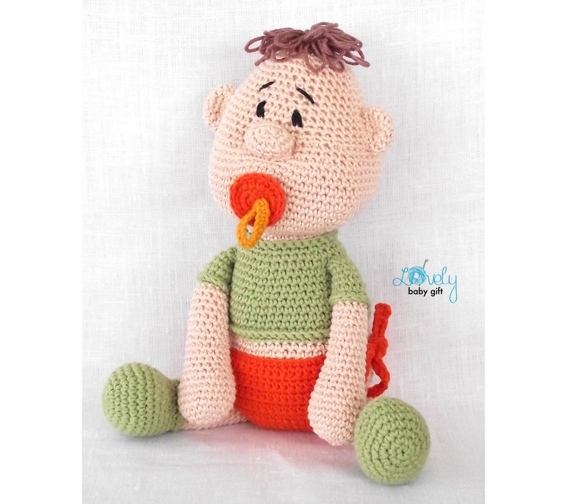 May include: A handmade crochet baby doll with a pacifier. The doll has a light skin tone, green shirt, orange shorts, and green feet. The doll's hair is brown, and it has a pacifier with orange and yellow accents. The doll is sitting.