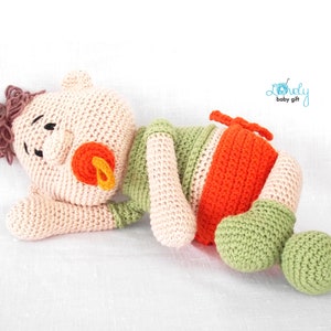 May include: A handmade crochet baby doll in a lying position. The doll features a light peach head with brown yarn hair, black eyes, and an orange pacifier. The body is green, orange, and peach. The text "Lovely baby gift" is in the upper right corner.