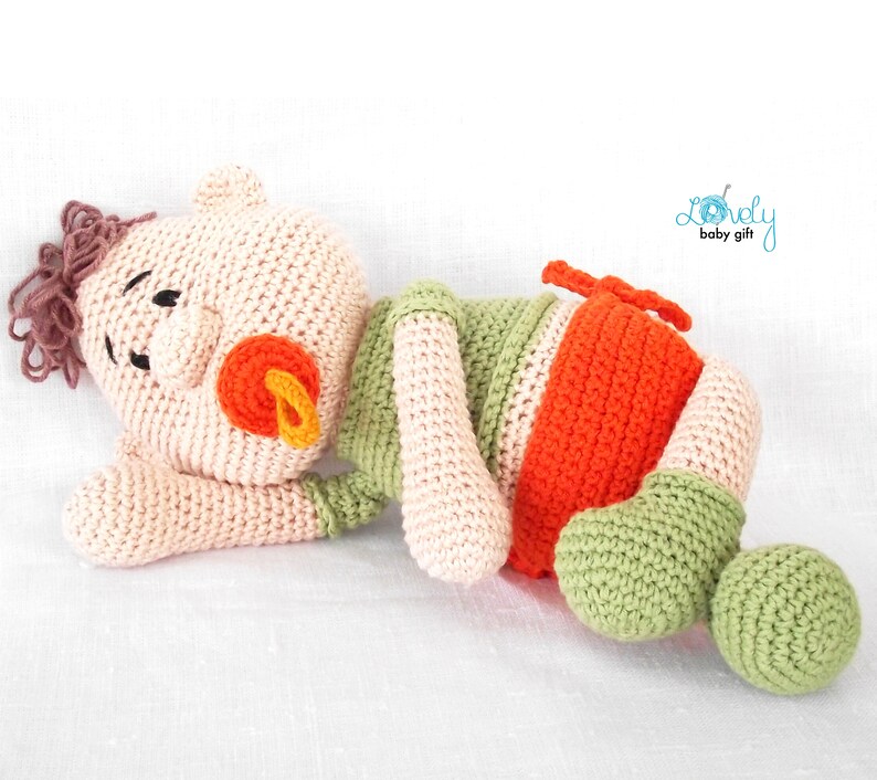 May include: A handmade crochet baby doll in a lying position. The doll features a light peach head with brown yarn hair, black eyes, and an orange pacifier. The body is green, orange, and peach. The text "Lovely baby gift" is in the upper right corner.