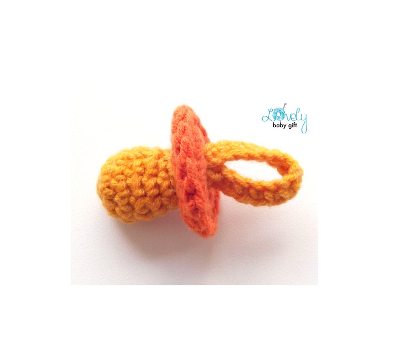 May include: A handmade crochet baby pacifier in shades of orange and yellow. The pacifier features a round nipple, a circular shield, and a loop for easy handling. The text "Lovely baby gift" is in the upper right corner.