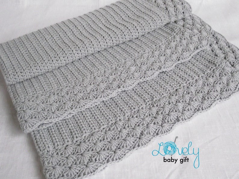May include: A gray crocheted baby blanket with a decorative border. The blanket is folded in thirds and has a subtle texture.