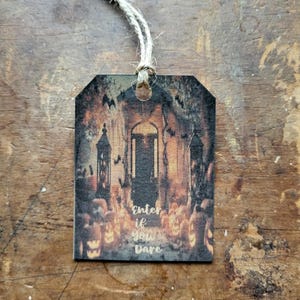 Puede incluir: Una etiqueta con temática de Halloween con una escena espeluznante que presenta una puerta, calabazas y murciélagos. La etiqueta tiene el texto "Enter if you Dare" y está unida a una cuerda de hilo natural. La etiqueta es de madera.