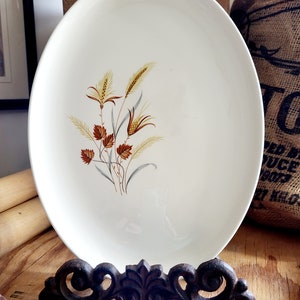 Puede incluir: Una fuente ovalada de color crema con un diseño de trigo y flores en tonos marrón, amarillo y gris. La fuente se exhibe sobre un soporte decorativo de metal oscuro. Una pieza vintage.