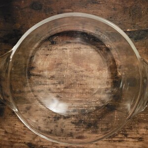 Puede incluir: Un molde para hornear de vidrio transparente con forma redonda y dos asas. El molde está vacío y se encuentra sobre una superficie de madera.