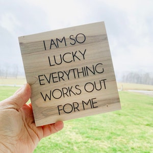 May include: Wooden sign with the text "I am so lucky everything works out for me" in black lettering on a light brown wood background.