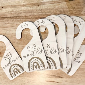 Puede incluir: Conjunto de etiquetas de percha de madera con diseños de arcoíris y texto que indica rangos de edad para recién nacidos hasta 24 meses. Las etiquetas incluyen "NB recién nacido", "0-3 meses", "3-6 meses", "6-9 meses", "9-12 meses", "12-18 meses" y "18-24 meses".