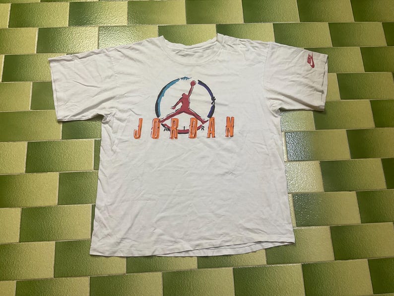 Puede incluir: Camiseta blanca con el logo de Jordan en rojo, naranja y azul. El logo muestra la silueta de un jugador de baloncesto en el aire, con la palabra "JORDAN" debajo. Peque&ntilde;o logo de Nike en la manga.