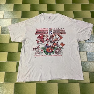 May include: White t-shirt featuring a graphic of a football game between the Nebraska Cornhuskers and the Virginia Tech Hokies. The graphic includes team mascots, helmets, and the text "DECEMBER 21ST, 1996 - PRO PLAYER STADIUM."