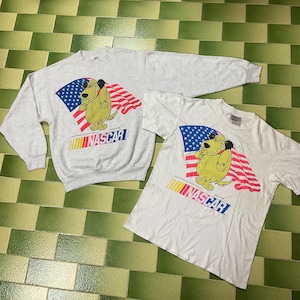 May include: A light gray sweatshirt and a white t-shirt featuring a cartoon dog with a flag design. The dog is yellow with black ears and is standing in front of a blue and red flag. The word "NASCAR" is printed below the dog.