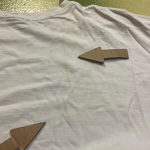 Puede incluir: Una camiseta blanca con dos flechas de cart&oacute;n marr&oacute;n que apuntan en diferentes direcciones. Las flechas son de cart&oacute;n corrugado y est&aacute;n colocadas en la parte delantera de la camiseta. La camiseta est&aacute; ligeramente arrugada.