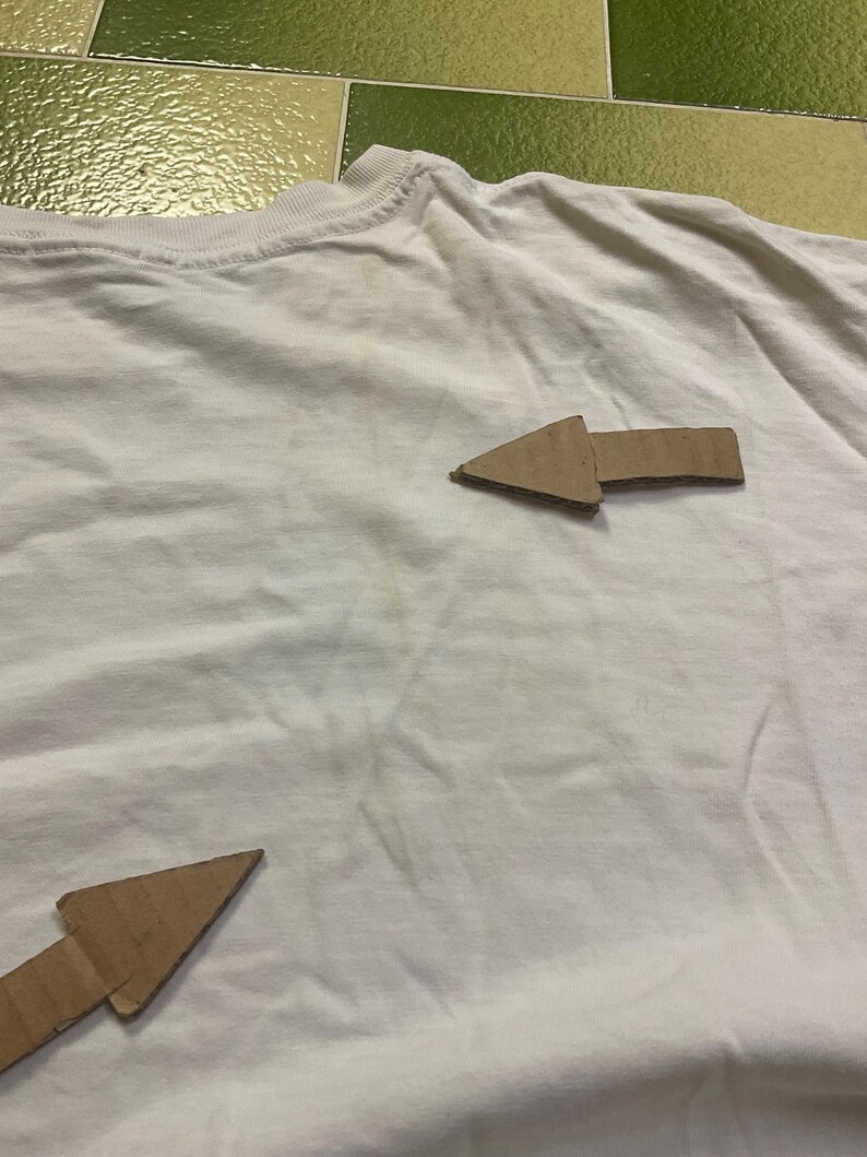 Puede incluir: Una camiseta blanca con dos flechas de cart&oacute;n marr&oacute;n que apuntan en diferentes direcciones. Las flechas son de cart&oacute;n corrugado y est&aacute;n colocadas en la parte delantera de la camiseta. La camiseta est&aacute; ligeramente arrugada.