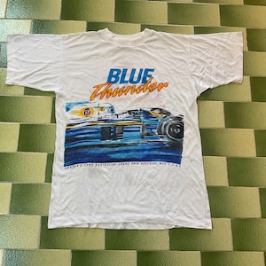 May include: White t-shirt featuring a racing car graphic with the words "BLUE Thunder" in blue and orange. The design includes the text "FOSTER'S 1989 AUSTRALIAN GRAND PRIX ADELAIDE, Nov. 2,3,4,5". The shirt is laid flat on a green and white tiled surface.