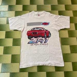 May include: White t-shirt featuring a red sports car graphic with the text "ZR1" in blue and silver. The shirt also includes the text "Life Begins at 180mph." The Chevrolet logo is above the car.