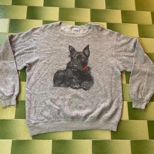 May include: A heather gray sweatshirt featuring a black illustration of a Scottish Terrier with a red collar. The long-sleeved crew neck top is made of a soft material.