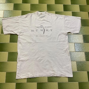 Op de afbeelding: Wit T-shirt met de tekst "THE MUMMY" in zwart, met een gestileerde mummie-afbeelding. Het shirt ligt plat, waarbij het ontwerp wordt getoond. Het T-shirt is gemaakt van een zacht materiaal.