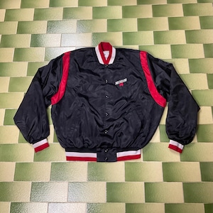 May include: Black satin bomber jacket with red and white accents. The jacket features a red collar and shoulder stripes, with white and red striped cuffs and hem. The word "Michelob" is embroidered on the chest.