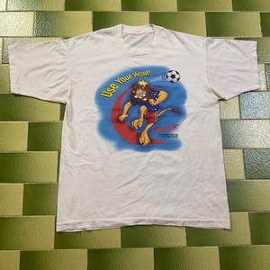 Puede incluir: Camiseta blanca con un león de dibujos animados pateando un balón de fútbol. El león lleva pantalones cortos y una camisa con estampado de estrellas. La camiseta tiene el texto "Use Your Head!" y "DARE".