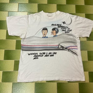 May include: White t-shirt with a cartoon illustration of two men in a cockpit. The shirt features text and a drawing of a jet. The text includes phrases like "Wapaya think I am like Maverick huh huh" and "Hen Hen Herben Do some of that pilot shit."
