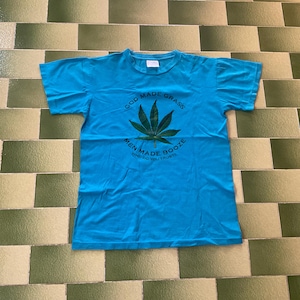 Puede incluir: Camiseta azul turquesa con un diseño de hoja de cannabis. El texto en la camiseta dice: "GOD MADE GRASS, MEN MADE BOOZE, WHO DO YOU TRUST?" La camiseta es de un material suave y es adecuada para uso casual.
