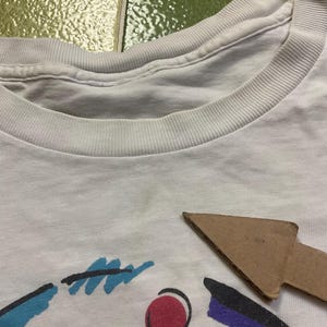 Puede incluir: Camiseta blanca con cuello redondo y dise&ntilde;o gr&aacute;fico colorido. El dise&ntilde;o incluye formas azules, rojas y moradas. La camiseta est&aacute; hecha de un material suave, probablemente algod&oacute;n, y parece vintage.