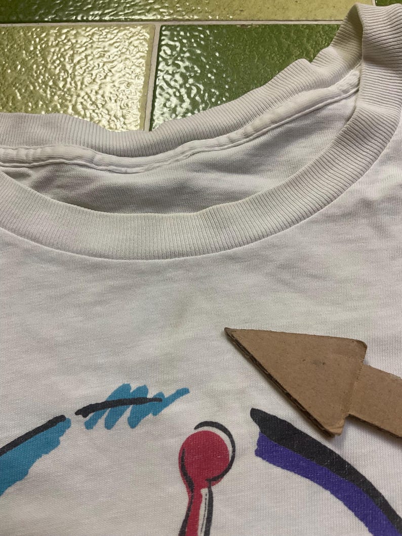 Puede incluir: Camiseta blanca con cuello redondo y dise&ntilde;o gr&aacute;fico colorido. El dise&ntilde;o incluye formas azules, rojas y moradas. La camiseta est&aacute; hecha de un material suave, probablemente algod&oacute;n, y parece vintage.