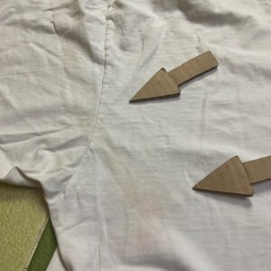 Puede incluir: Una camiseta blanca con cuello redondo, que muestra signos de desgaste. Dos flechas de cart&oacute;n apuntan hacia la prenda. La tela est&aacute; arrugada y tiene manchas.