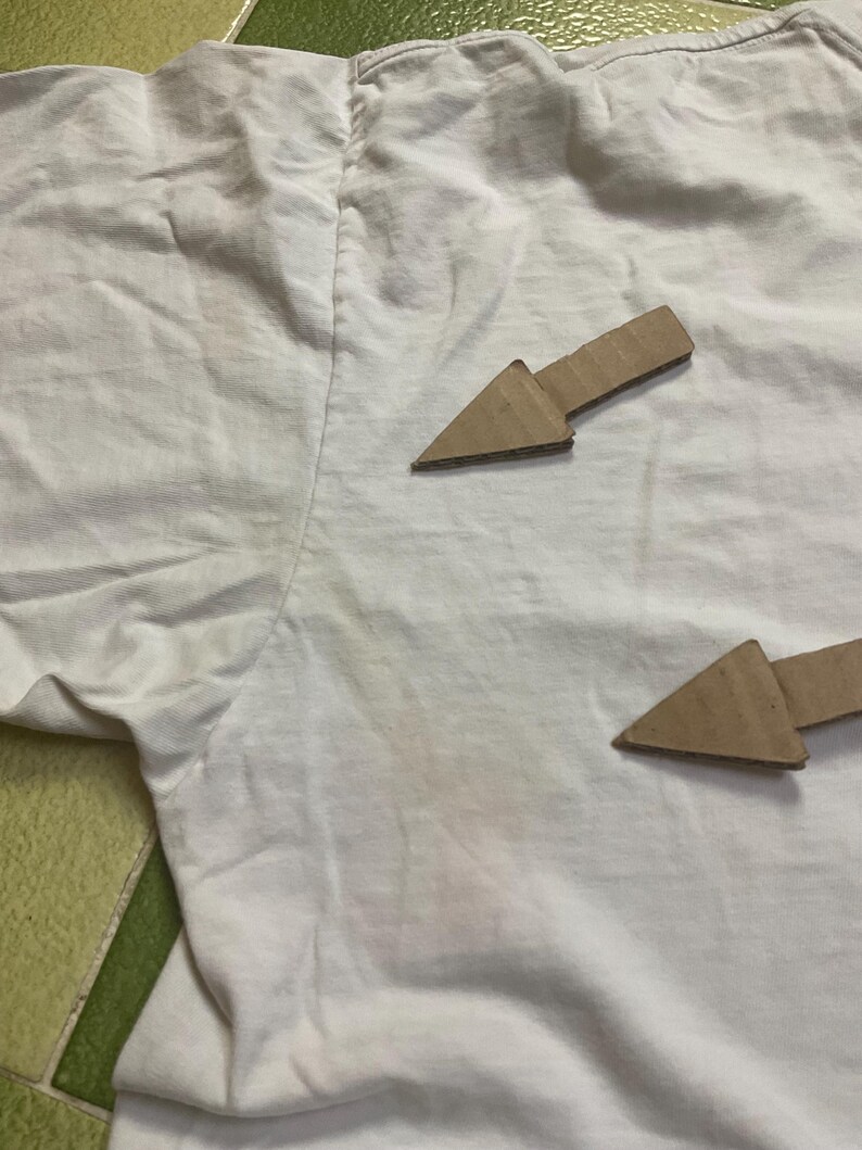 Puede incluir: Una camiseta blanca con cuello redondo, que muestra signos de desgaste. Dos flechas de cart&oacute;n apuntan hacia la prenda. La tela est&aacute; arrugada y tiene manchas.