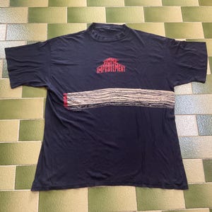 Puede incluir: Camiseta azul marino con el texto rojo "HOME IMPROVEMENT" sobre un diseño horizontal beige y negro. La camiseta tiene mangas cortas y cuello redondo.