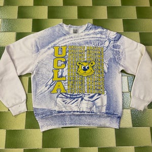 Könnte beinhalten: Weißes Sweatshirt mit blau-gelber UCLA Bruins Grafik. Das Design zeigt das Wort "UCLA" in gelben Blockbuchstaben, einen Cartoon-Bären und das Wort "BRUINS" vertikal wiederholt. Die Ärmel sind einfarbig weiß.
