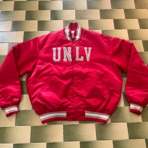 Könnte beinhalten: Rote Satin-Collegejacke mit den Buchstaben "UNLV" in Weiß auf der Brust. Die Jacke hat einen weißen Kragen und Manschetten mit grauen und roten Streifen. Ein klassischer College-Stil.