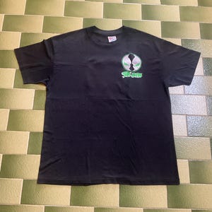 Op de afbeelding: Een zwart t-shirt met korte mouwen en een cirkelvormige afbeelding op de linkerborst. De afbeelding toont een wit en groen maskerachtig embleem met het woord "SPAWN" in groene letters direct onder het cirkelvormige ontwerp.