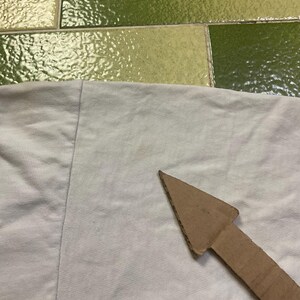 Puede incluir: Primer plano de una tela de color claro con una flecha de cart&oacute;n que apunta hacia la derecha. La tela parece ser de color gris claro o blanquecino. La flecha es de cart&oacute;n marr&oacute;n.