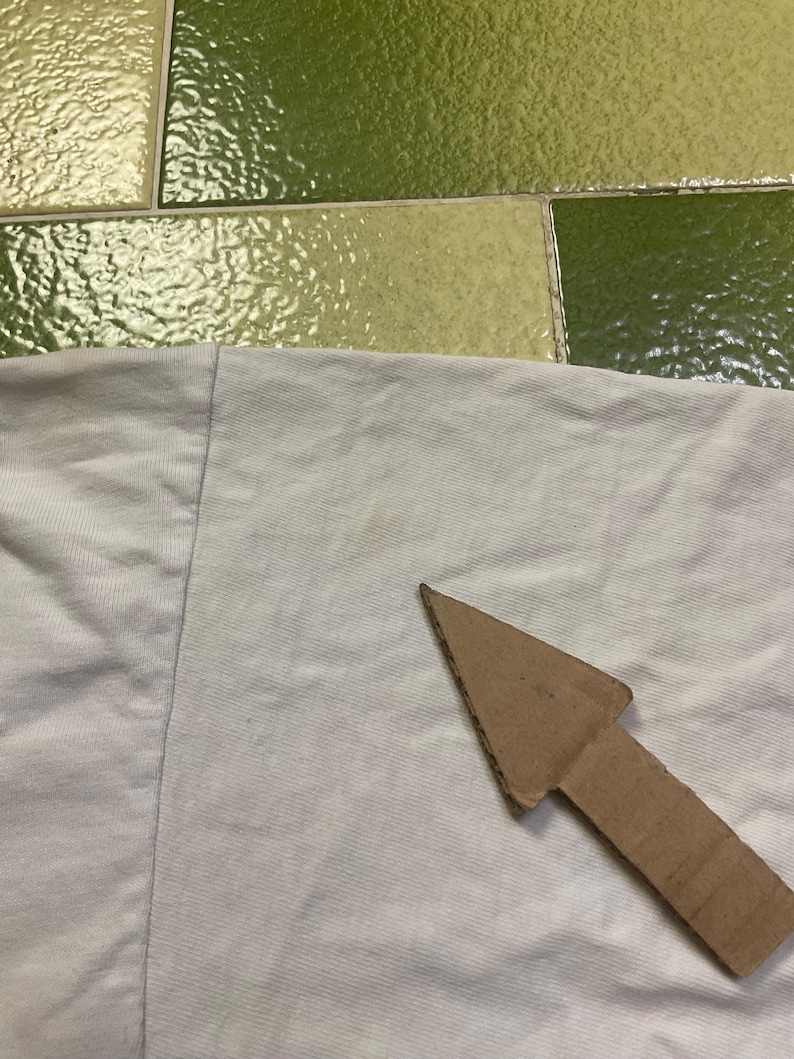 Puede incluir: Primer plano de una tela de color claro con una flecha de cart&oacute;n que apunta hacia la derecha. La tela parece ser de color gris claro o blanquecino. La flecha es de cart&oacute;n marr&oacute;n.