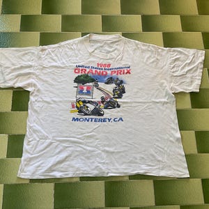 May include: White t-shirt featuring a colorful graphic of motorcycles racing on a track. The text on the shirt reads "1988 United States International Grand Prix" and "Monterey, CA."