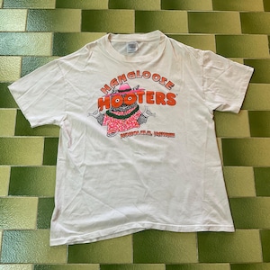 May include: White t-shirt featuring the text "HANGLOOSE HOOTERS" in orange and a cartoon character wearing a pink hat and shirt. The text "HONOLULU, HAWAII" is printed below the graphic. The shirt is short-sleeved.