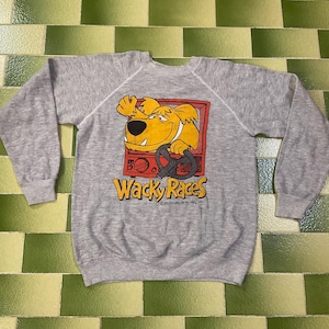 May include: A gray sweatshirt featuring a cartoon dog from Wacky Races. The dog is yellow and appears to be driving a vehicle. The words "Wacky Races" are printed in yellow and red below the image.