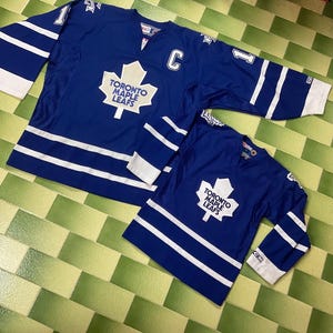 May include: Two blue hockey jerseys with white stripes and accents. The jerseys feature the Toronto Maple Leafs logo and the letter "C". One jersey is larger than the other, suggesting a parent and child set. The jerseys are on a green and yellow tiled surface.